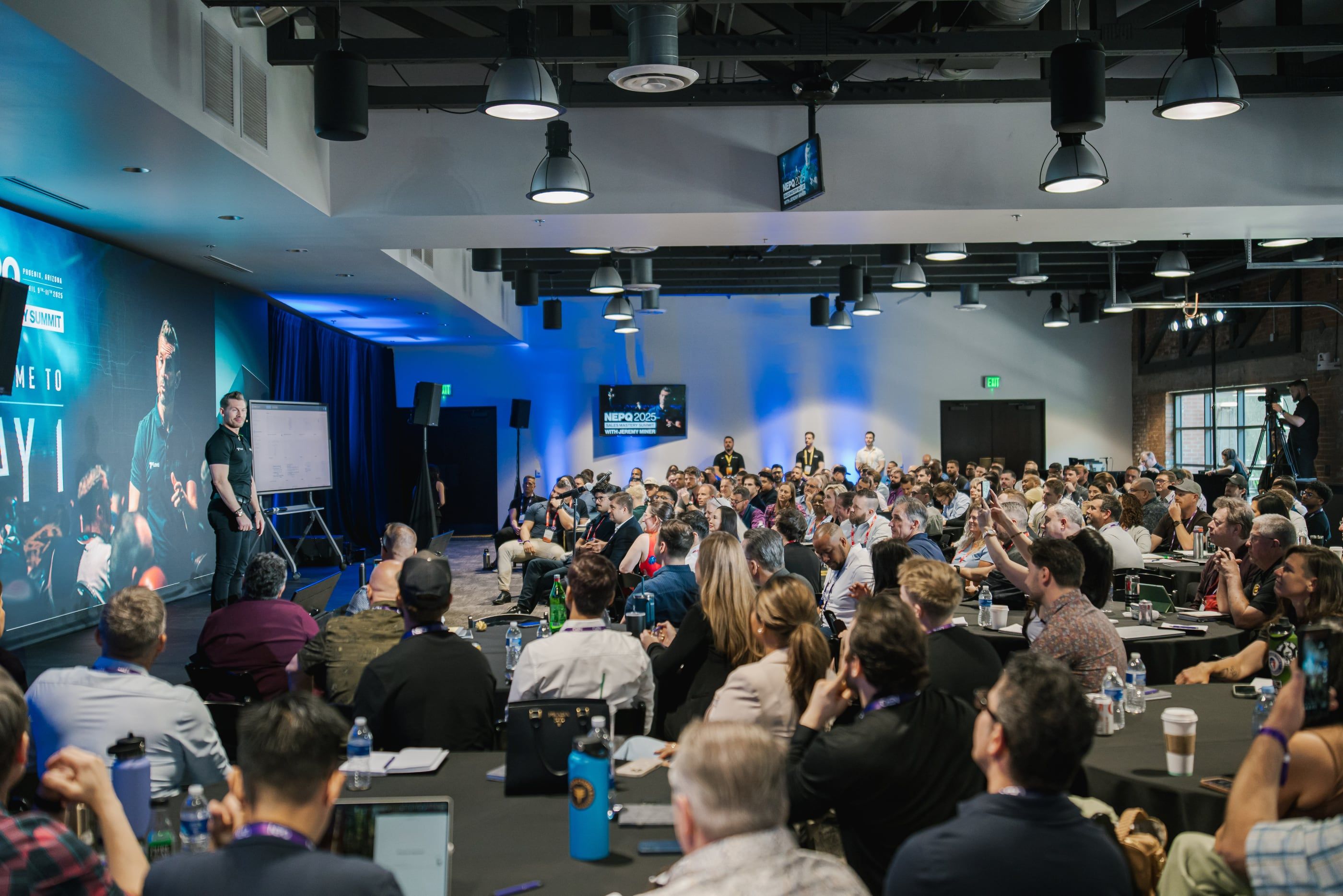 The summit room filled with attendees at round tables while Jeremy Miner presents on stage